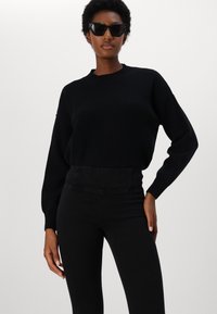 Suéter negro de gran tamaño con textura acanalada y cuello redondo, combinado con jeans negros de cintura alta. La modelo lleva gafas de sol negras y angulares.