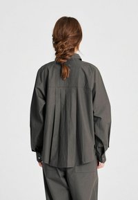 Camicia a maniche lunghe in tessuto grigio scuro con vestibilità rilassata, caratterizzata da pieghe lungo la schiena e polsini con bottoni. Texture liscia e design semplice.
