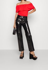 Haut rouge décolleté sur les épaules, pantalon noir brillant taille haute et talons à plateforme noirs. Mannequin tenant un petit sac à main noir.