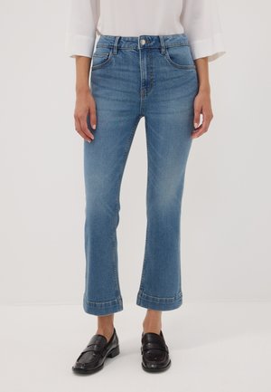 Mujer con vaqueros azules recortados de pierna recta, blusa blanca y mocasines negros, de pie contra un fondo liso.