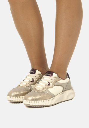 Sneakers con una toma in tessuto metallico dorato e parte superiore in pelle bianca, caratterizzati da una suola bianca textured, dettagli viola e un design con lacci.