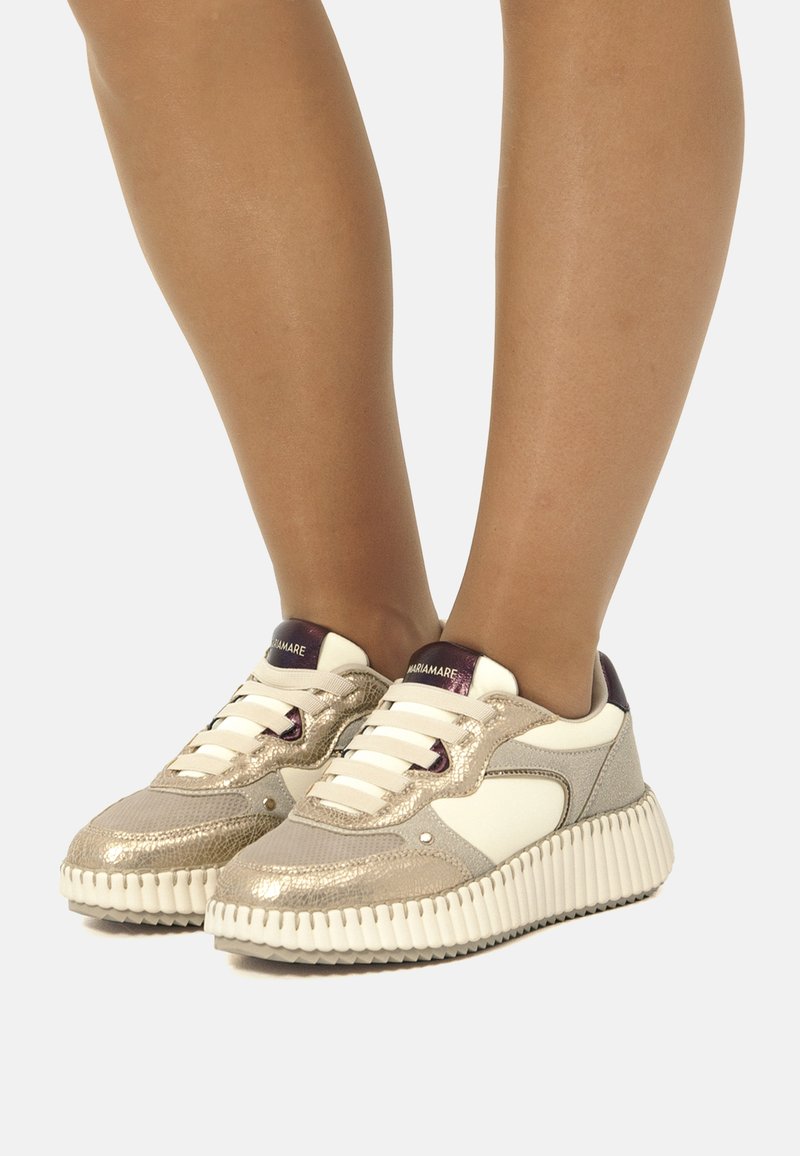Zapatillas con parte superior de textil metálico dorado y cuero blanco, que cuentan con una suela texturizada blanca, acentos morados y un diseño de cordones.