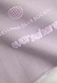 Gros plan sur un tissu lavande avec le texte en relief « WELLNESS CLUB SOCIETY » et « everywhere » ainsi qu'une icône de globe.