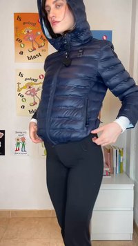 Femme portant une doudoune marine à capuche et un pantalon noir ajustant sa veste dans une pièce avec des affiches colorées et une étagère blanche.