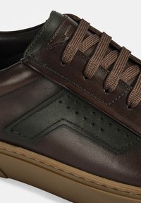 Braune Ledersneaker mit grünen Akzenten, ausgestattet mit strukturiertem Material, dünnen braunen Schnürsenkeln und perforierten Details an der Seite. Gummisohle.
