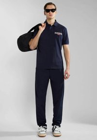 Tummansininen polo-paita, jossa on logo, yhdistettynä tummiin housuihin. Malli pitää mustaa urheilulaukkua, käyttää aurinkolaseja ja valkoisia lenkkareita, joissa on värikohokuvioita.