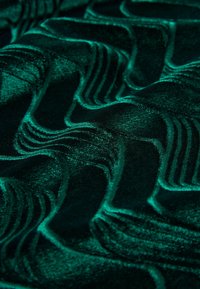 Teksturowana tkanina w kolorze szmaragdowej zieleni z falistym wzorem, charakteryzująca się miękką, puchową powierzchnią i subtelnym połyskiem, wyeksponowująca skomplikowane detale wzoru.