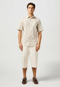 Camicia a maniche corte beige a righe con chiusura a bottoni, abbinata a pantaloni bianchi tagliati e scarpe da barca marroni.