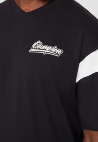 Camiseta de algodón negra con cuello en V, con un panel blanco en el hombro y un logo blanco de "Champion" en el lado izquierdo del pecho. Textura suave, diseño casual.