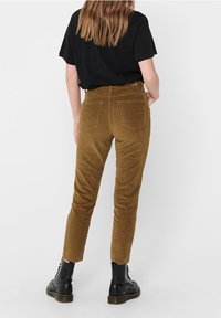 Pantalon en velours côtelé marron coupe slim, avec deux poches arrière et ourlets effilochés, porté avec une chemise noire et des bottes noires à lacets.