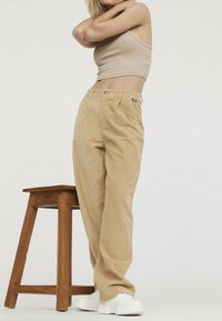 Pantalons en velours côtelé beige avec une coupe large, dotés de deux poches avant et d'un discret côtelage vertical. Associés à des baskets blanches.