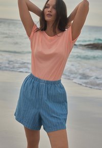 Kerge roosa lühikeste varrukatega t-särk, mis on kombineeritud siniste triibuliste lühikesed püksidega. T-särk on pehme, samas kui püksidel on elastne vöökoht ja küljetaskud.