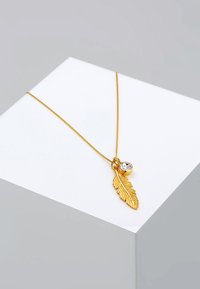 Goldfarbenes Halsband mit einem Blattanhänger, der strukturierte Details und einen runden Kristallakzent aufweist, präsentiert auf einem weißen Sockel.