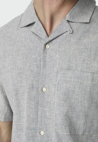 Närbild av en person som bär en kortärmad, ljusgrå skjorta med tunna vertikala vita ränder, en bröstficka och en spetsig krage.