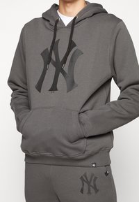 Sweat à capuche gris en mélange de coton, avec un logo noir sur la poitrine, une capuche ajustable à cordon et une poche kangourou. Poignets et ourlet côtelés.