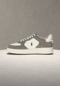 Λευκό και γκρι χαμηλό sneaker με κορδόνια στο μπροστινό μέρος και λογότυπο Polo στο πλάι και στη φτέρνα, εμφανίζεται σε φωτεινή επιφάνεια με γκρι φόντο.