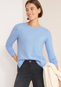 Maglione azzurro chiaro in morbido filato con scollatura rotonda, polsini a coste e dettagli a treccia sulle maniche, abbinato a jeans neri.