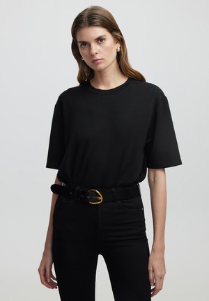 Schwarzes Baumwoll-T-Shirt mit Rundhalsausschnitt und kurzen Ärmeln, kombiniert mit hochgeschnittenen schwarzen Jeans und einem schwarzen Gürtel mit einer goldenen runden Schnalle.