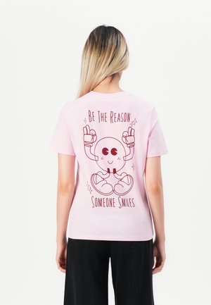 Personne avec de longs cheveux portant un t-shirt rose sur lequel est représenté un personnage de dessin animé et le texte "Soyez la raison pour laquelle quelqu'un sourit" au dos.