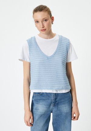 Chaleco de punto azul claro con cuello en V y diseño sin mangas, superpuesto a una camiseta blanca, combinado con jeans azules. Patrón texturizado en toda la prenda.