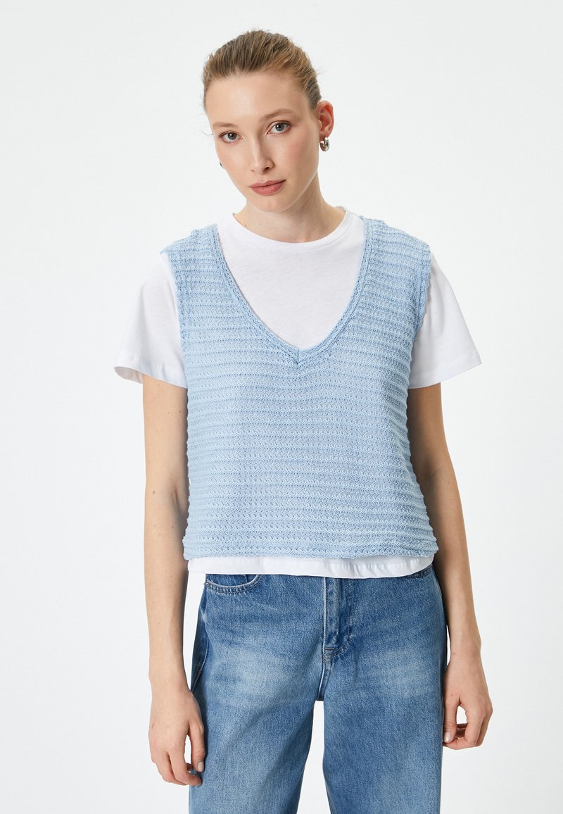 Chaleco de punto azul claro con cuello en V y diseño sin mangas, superpuesto a una camiseta blanca, combinado con jeans azules. Patrón texturizado en toda la prenda.