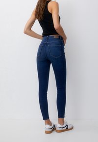 Jean skinny en denim brut foncé avec une taille haute. Poches arrière et texture lisse. Associé à un haut sans manches noir et des baskets blanches.