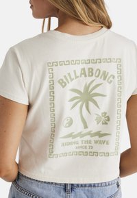 Cremefarvet bomulds crop top med et grønt grafisk design af et palmetræ, yin-yang og teksten "Riding the Wave since 73" på bagsiden.