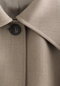 Manteau en tissu beige texturé avec large col et grand bouton noir à quatre trous fermé près de l'encolure.