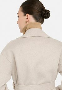 Cappotto beige in misto lana con colletto sciallato, vita con cintura e texture morbida, indossato sopra un maglione a collo alto marrone chiaro. Orecchini a cerchio dorati inclusi.