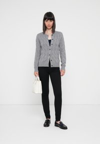 Cardigan grigio lavorato a maglia con motivo a trecce, scollatura rotonda e chiusura con bottoni. Abbinato a pantaloni neri e mocassini neri, mentre tiene una borsa bianca.