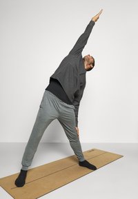 Homme en sweat à capuche gris foncé et pantalon gris clair s'étirant sur le côté sur un tapis de yoga marron dans une pièce lumineuse.