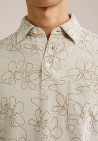 Camisa de color beige claro con un patrón floral en marrón, fabricada en un tejido texturizado, con un cuello con cierre de botón y un bolsillo en el pecho izquierdo.