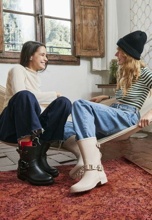 Černá gumová kotníková bota s detaily na sponě a červeným akcentem; světle zbarvená gumová bota se sponou; obě boty jsou umístěny na texturovaném koberci.