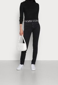 Svarta högmidjade jeans med vitt sidologotypdetalj, ihopparade med en figurnära svart polotröja och en vit vadderad handväska. Vita sneakers.