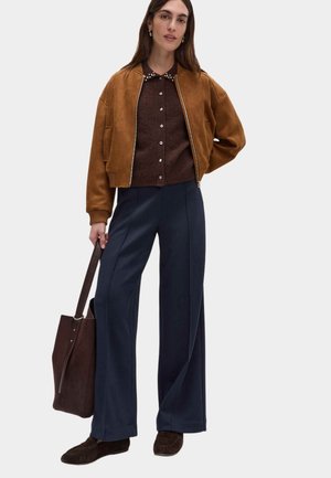 Kvinde iført brun ruskindsbomberjakke, mørkebrun knappet sweater, marineblå viddebenbukser, brune mokkasiner, holder mørkebrun tote taske.