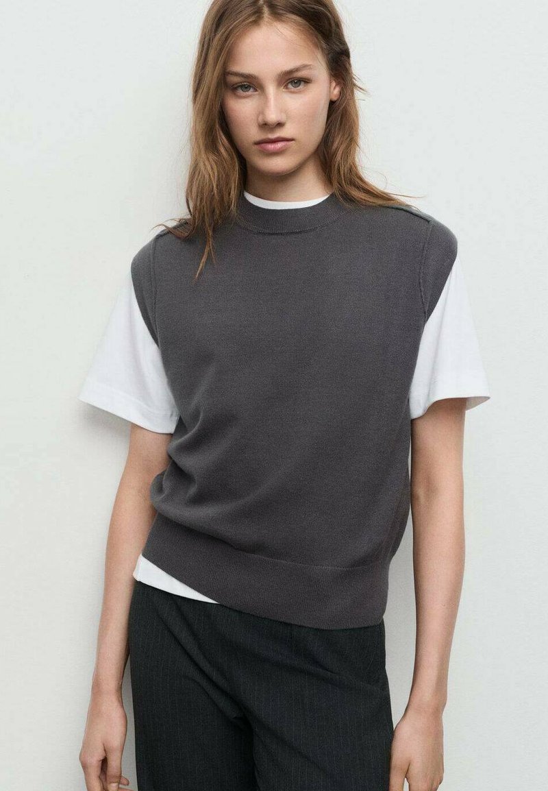 Grå kortärmad tröja över en vit t-shirt, ribbad halsringning och nederkant. Mjuk textur, avslappnad passform, i kombination med mörka randiga byxor.
