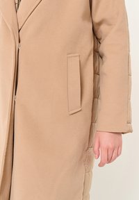 Cappotto lungo beige con una texture liscia, tasca laterale e dettagli trapuntati su un lato. Chiusura con zip visibile.