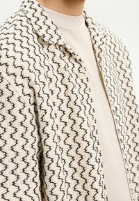 Vävd beige jacka med svart zigzag-mönster, med krage och knappstängning. Texturerad tyg ger visuellt intresse.