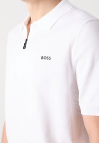 Polo blanc à manches courtes, col zippé et logo discret. Présente des accents en mesh texturé sur les côtés pour la respirabilité.