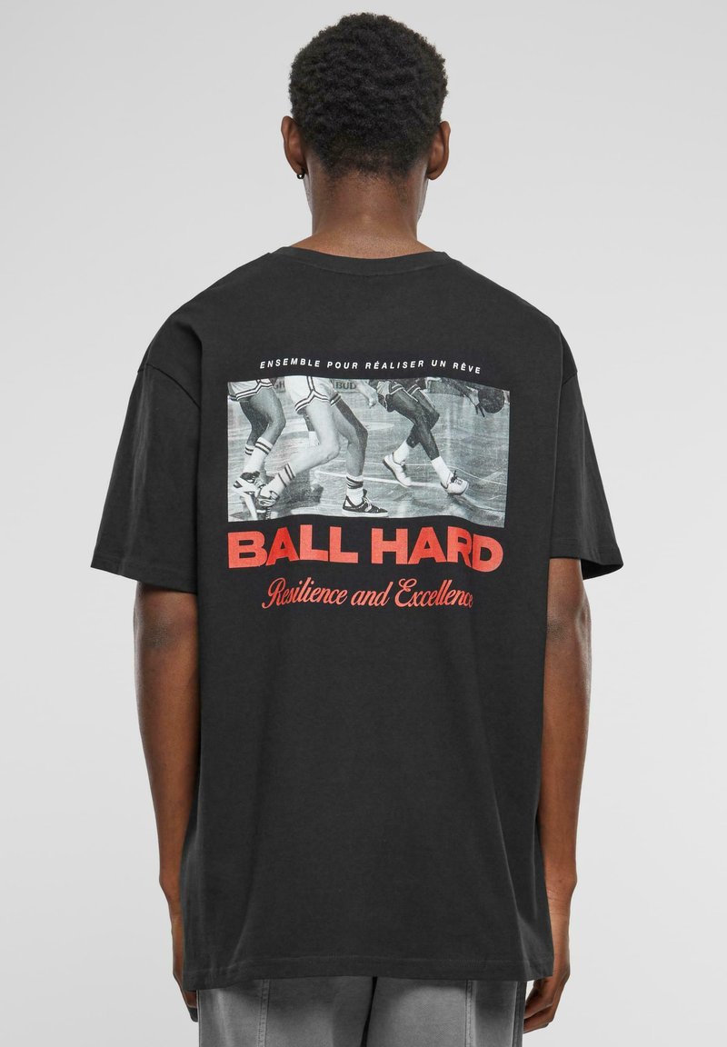 Sort bomulds t-shirt med et grafisk print, der viser sneakers og rød tekst "BALL HARD Resilience and Excellence" på ryggen. Korte ærmer.
