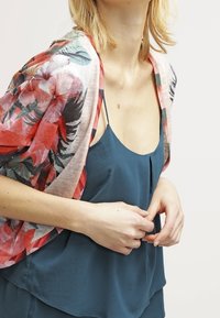 Blumenkimono aus durchscheinendem pinkfarbenem Stoff mit roten und grünen Mustern, über einem blauen ärmellosen Oberteil mit einer dezenten Faltenoptik getragen.