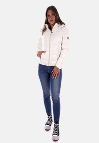 Giacca invernale bianca con zip, colletto alto e tasche laterali, abbinata a jeans skinny blu e sneakers nere alte.