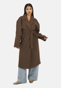 Cappotto in misto lana marrone con colletto a revers, maniche lunghe e vita fasciata. Presenta tasche laterali e un fit rilassato. Abbinato a jeans a gamba larga.