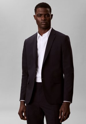 Mann trägt einen dunklen Anzug mit weißem Hemd, steht vor einem einfarbigen hellgrauen Hintergrund und blickt nach vorne.