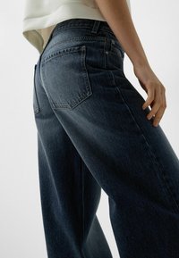Vi-ser ett par vid ben dark blue denimjeans med hög midja, med bakfickor och en slät textur, framhävd av subtila blekningar.