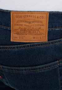 Jean en denim bleu foncé de Levi's, avec une pièce en cuir marron clair à l'arrière comportant un logo embossé, taille Lot 512 W31 L32, et des coutures jaunes.