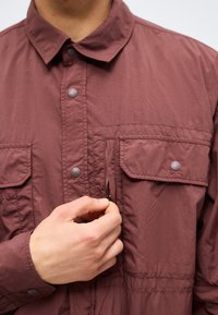Person wearing a maroon button-up shirt, using right hand to unzip the vertical chest pocket on the left side of the shirt.