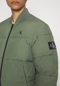 Groene gewatteerde jas van synthetisch materiaal, met een zwarte ritssluiting, geribde kraag en een geborduurd logopatch op de mouw.