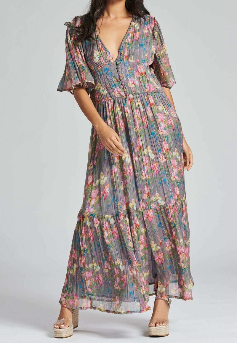Maxi-robe fleurie grise avec une texture plissée. Comprend des manches courtes à volants, un décolleté en V profond et un ourlet à volants. Portée avec des sandales compensées.