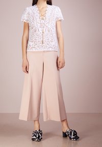 Kobieta ubrana w biały koronkowy top z krótkim rękawem, różowe szerokie spodnie o długości 7/8, granatowe kwiatowe klapki na obcasie oraz długi złoty naszyjnik.